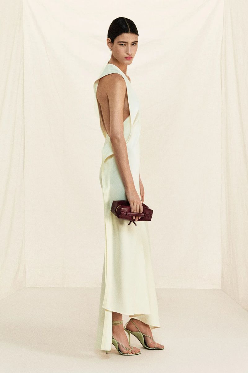 Fashion shot of a female model in a pale yellow gown with a burgundy clutch and gold heels against a neutral studio backdrop.