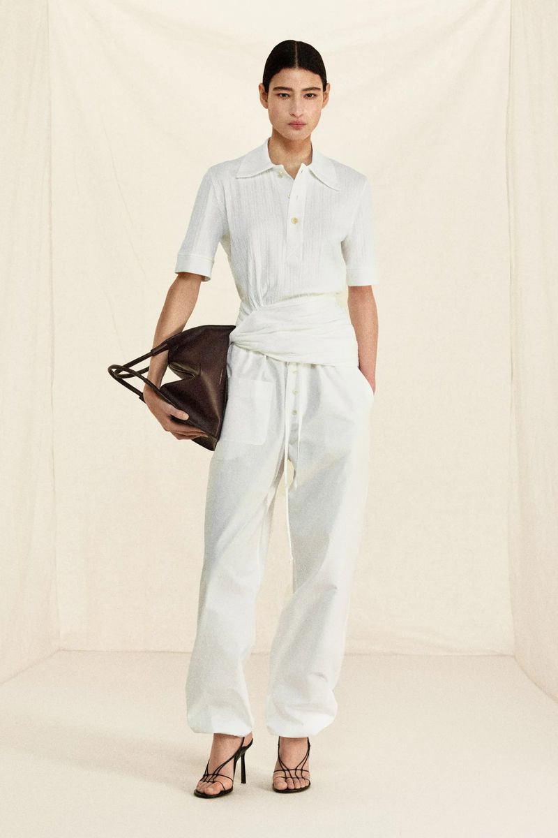 Woman in white shirt and trousers holding a brown bag, posing in front of a plain white backdrop, fashion studio shot.