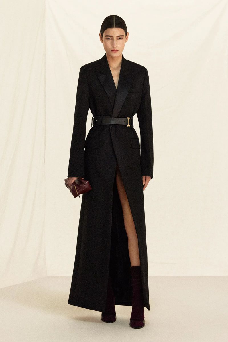 Fashionable woman in a long black jacket-dress with a belt, holding a burgundy clutch, posing against a neutral backdrop in a studio.