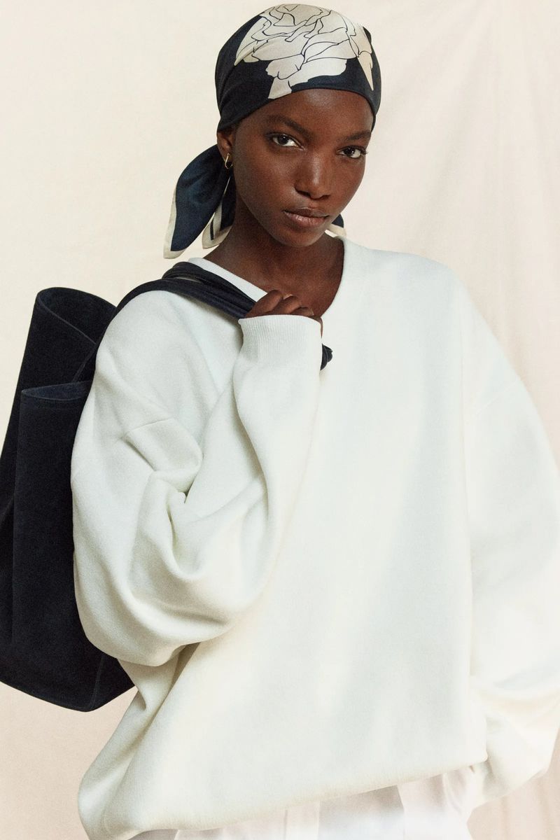 A dark-skinned female model poses indoors wearing a white sweatshirt and a patterned headscarf, with a black bag slung over her shoulder.