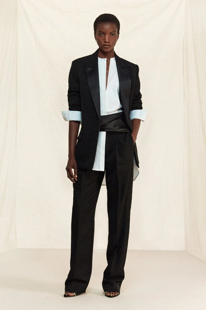 A fashion shot of a Black woman in a black tuxedo suit with a light blue shirt and satin cummerbund, posing against a neutral backdrop.