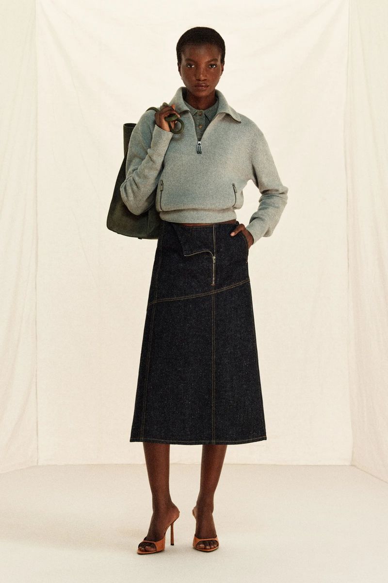 Full shot of a dark-skinned woman in a grey zip-up fleece, denim skirt, and heels, carrying a tote bag in a neutral indoor space.