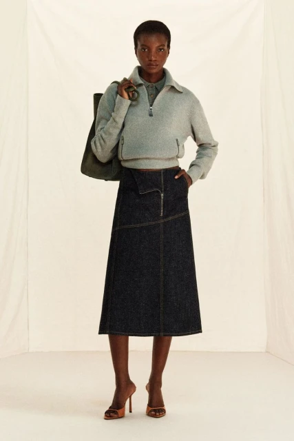 Full shot of a dark-skinned woman in a grey zip-up fleece, denim skirt, and heels, carrying a tote bag in a neutral indoor space.