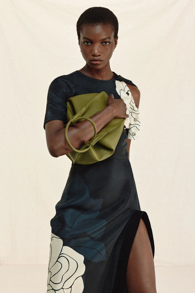 Elegant woman poses in a black floral print dress with a high slit, holding a green ring bag against a light background in a fashion editorial.