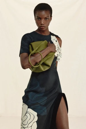 Elegant woman poses in a black floral print dress with a high slit, holding a green ring bag against a light background in a fashion editorial.