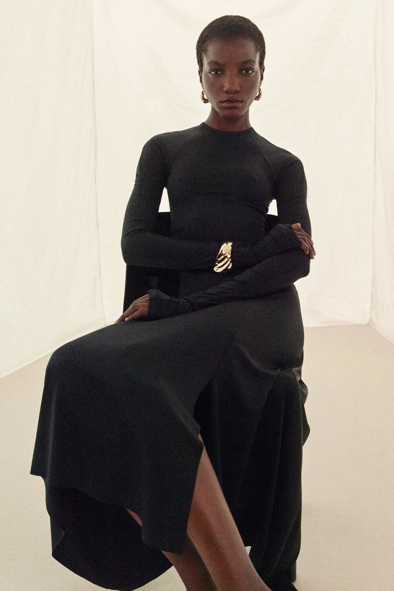 Elegant Black Woman in Long-Sleeve Dress Posing Seated with Bracelet. Fashion portrait in bright, neutral studio setting with soft light.