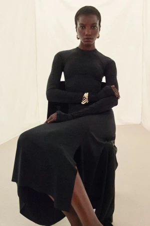 Elegant Black Woman in Long-Sleeve Dress Posing Seated with Bracelet. Fashion portrait in bright, neutral studio setting with soft light.