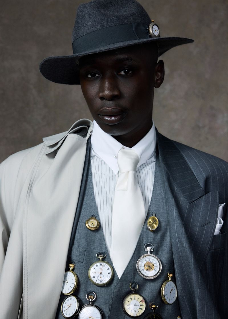 Stylish portrait of an adult male wearing a hat and suit vest covered in pocket watches in a photo studio setting.