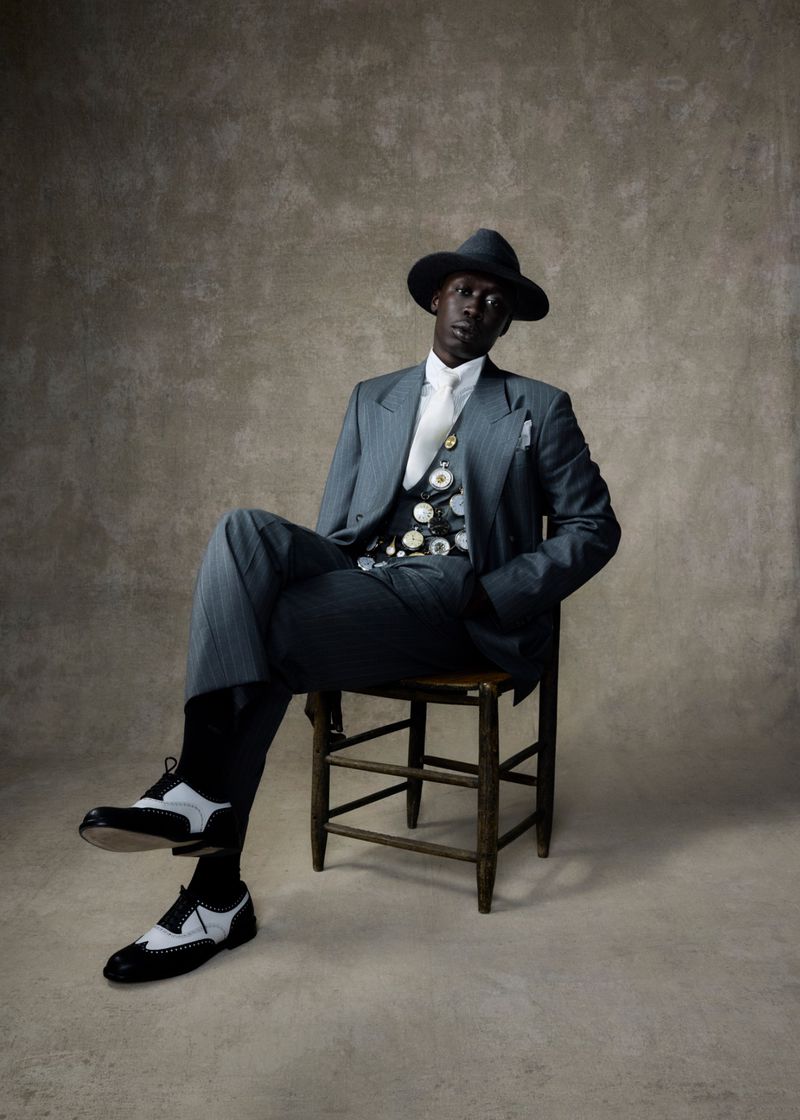 Stylish African American man in a gray suit, hat, and spectator shoes seated on a wooden chair against a mottled neutral backdrop.