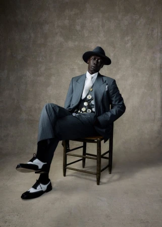 Stylish African American man in a gray suit, hat, and spectator shoes seated on a wooden chair against a mottled neutral backdrop.