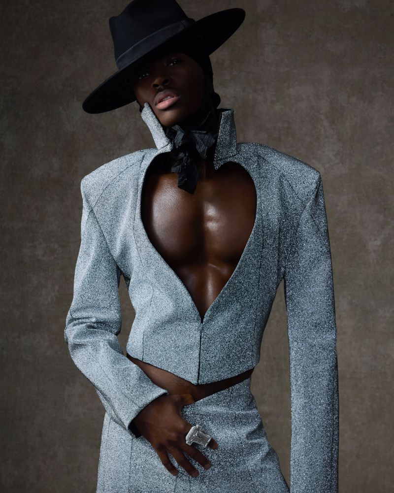 A dark-skinned adult with a black hat and sparkling gray jacket poses against a brown backdrop indoors, showcasing fashion and style.