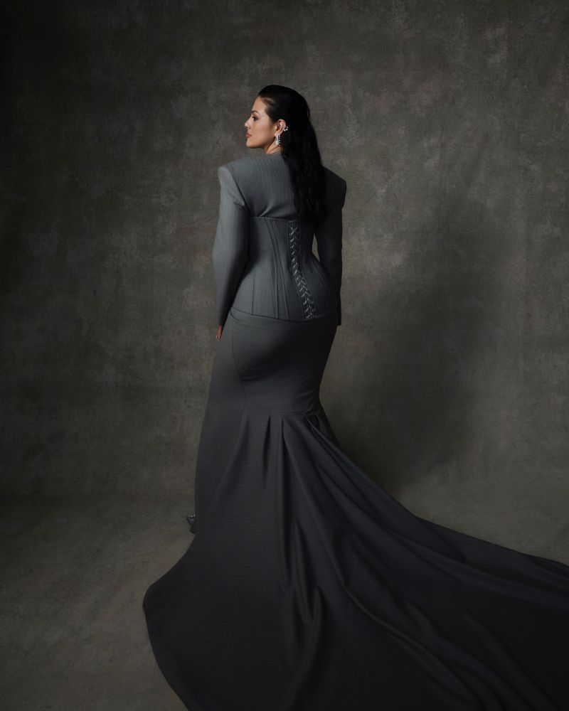 A woman poses in a grey jacket, corset, and gown with a long train against a mottled grey backdrop in a studio setting.