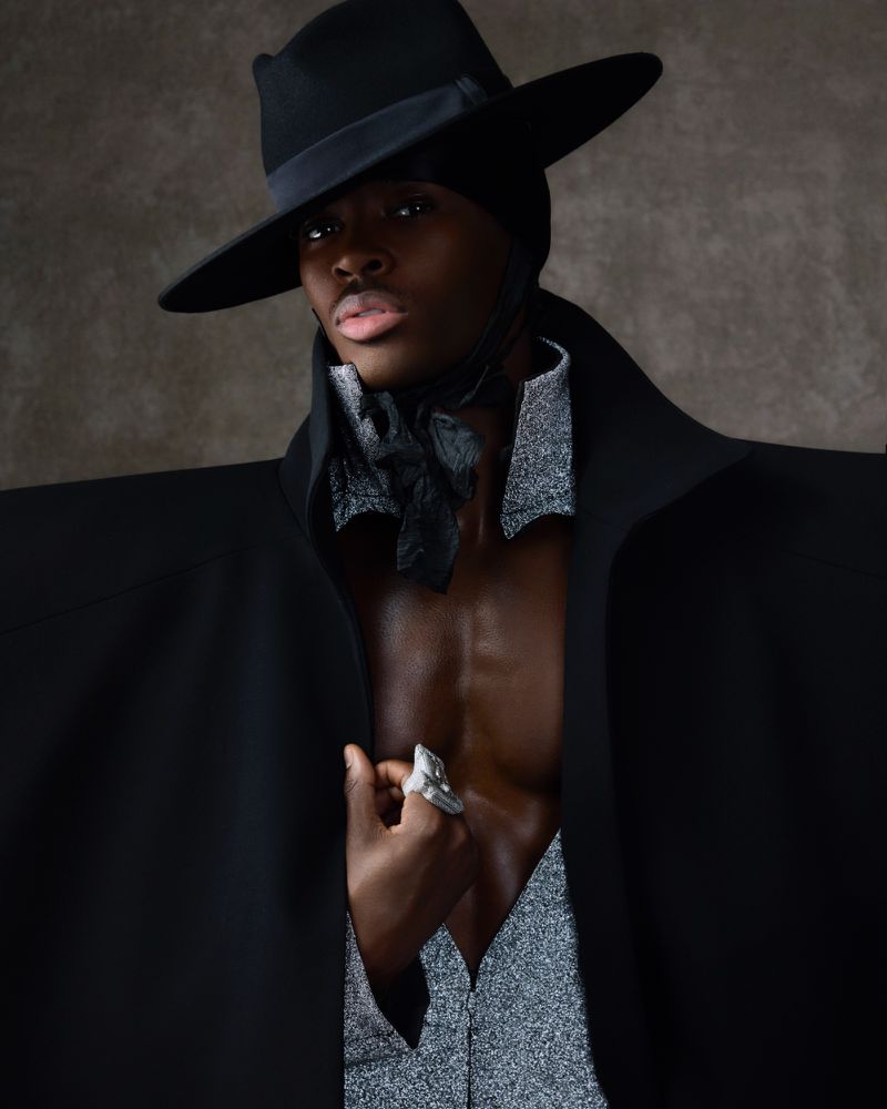 Person wearing a black hat and coat with a silver shirt and ring, posing in front of a mottled grey background in a studio setting.