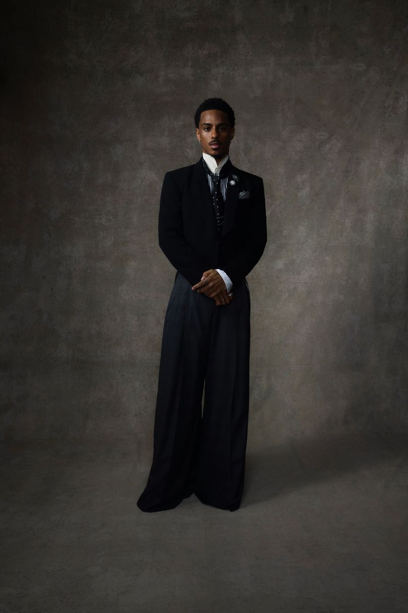 Portrait of an adult male in formal attire standing against a textured grey backdrop in a studio setting, lit with diffused lighting.