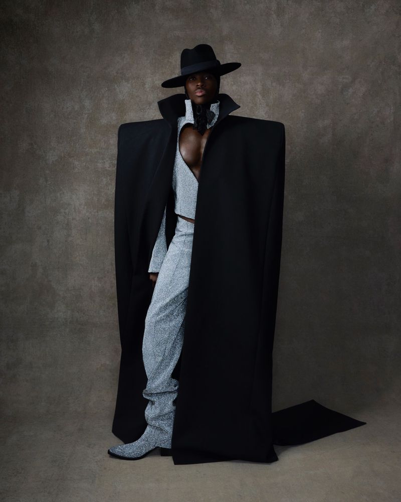 Fashionable adult with dark skin in a grey sequined outfit and black hat and cape poses in a studio with a neutral-toned background.