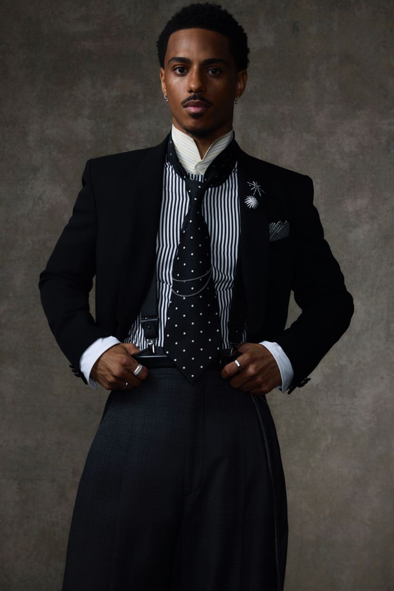Portrait of an adult male in a black suit and dotted tie against a textured gray backdrop, striking a confident pose.