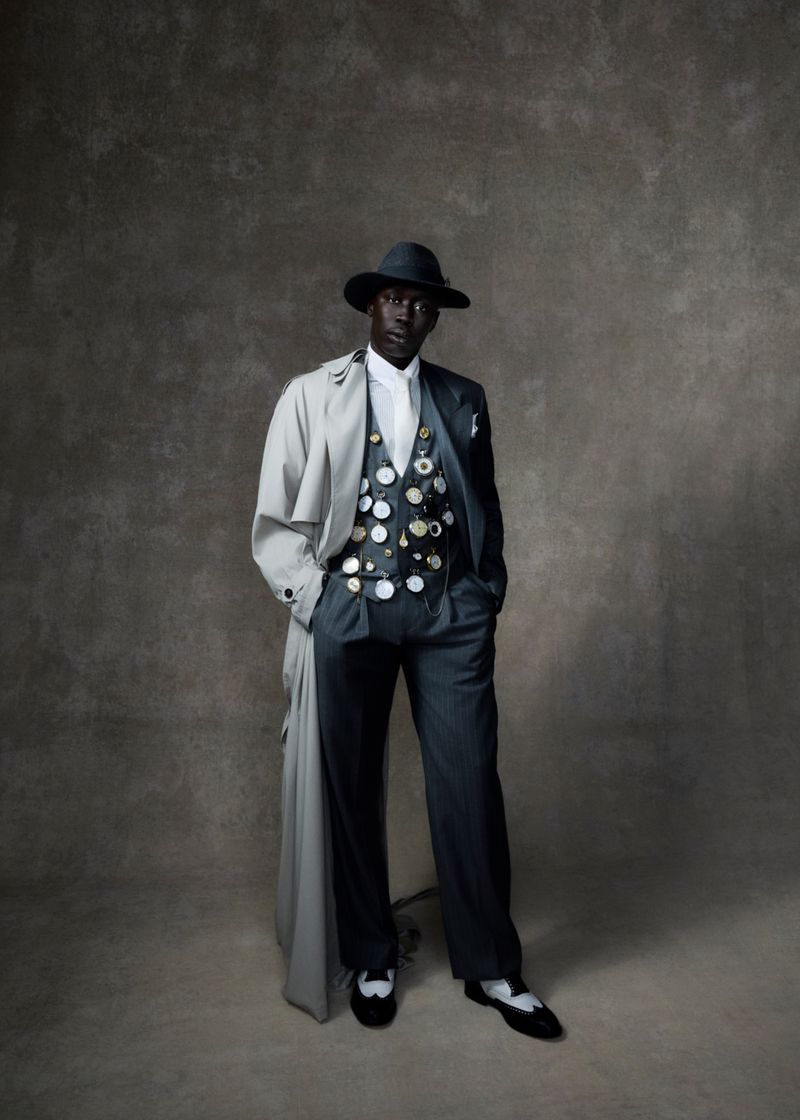 Full-length portrait of Black male in a hat, light trench coat, vest with buttons, and pinstripe pants in front of a mottled grey background.