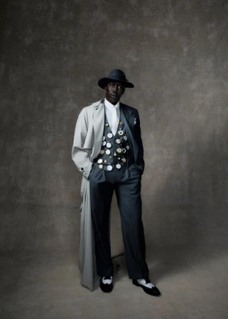 Full-length portrait of Black male in a hat, light trench coat, vest with buttons, and pinstripe pants in front of a mottled grey background.