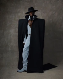 Model wearing an oversized black coat with structured shoulders, wide-brim hat, and patterned trousers against a mottled dark background.