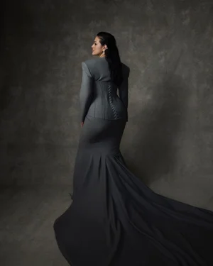 Ashley Graham wearing a grey tailored gown with a corset back and long train against a textured grey background.