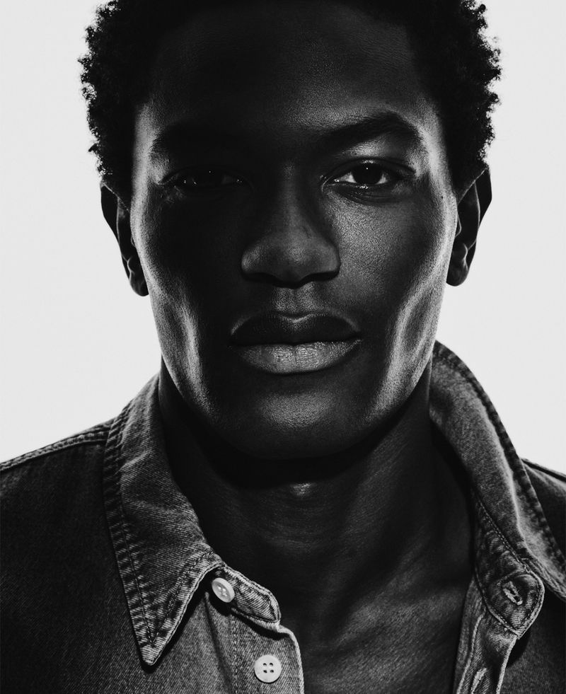 Striking portrait of a dark-skinned adult male wearing a denim shirt, captured in black and white with dramatic lighting.