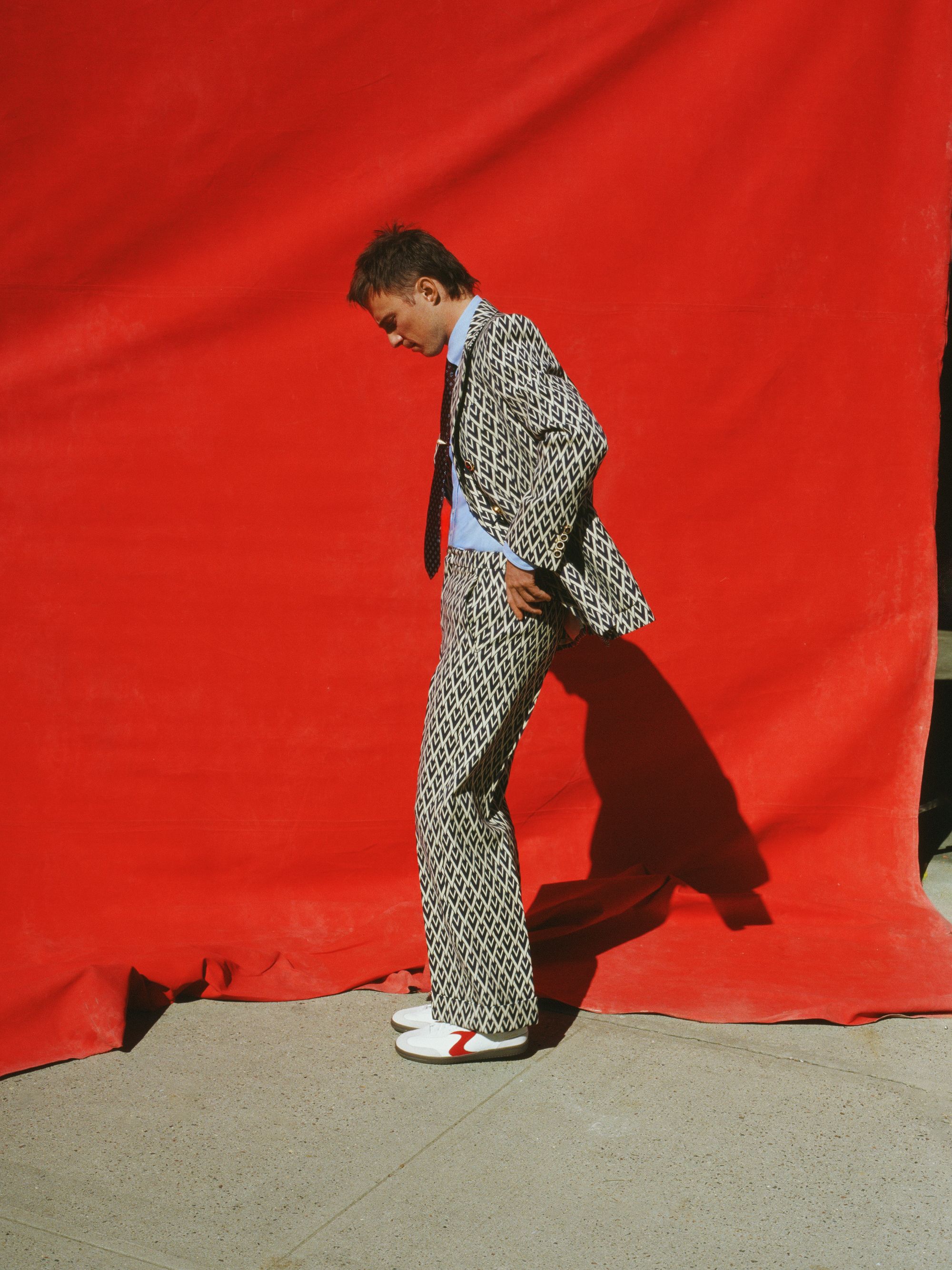 Young man in patterned suit stands confidently against a vibrant red backdrop, showcasing a blend of vintage and contemporary fashion.