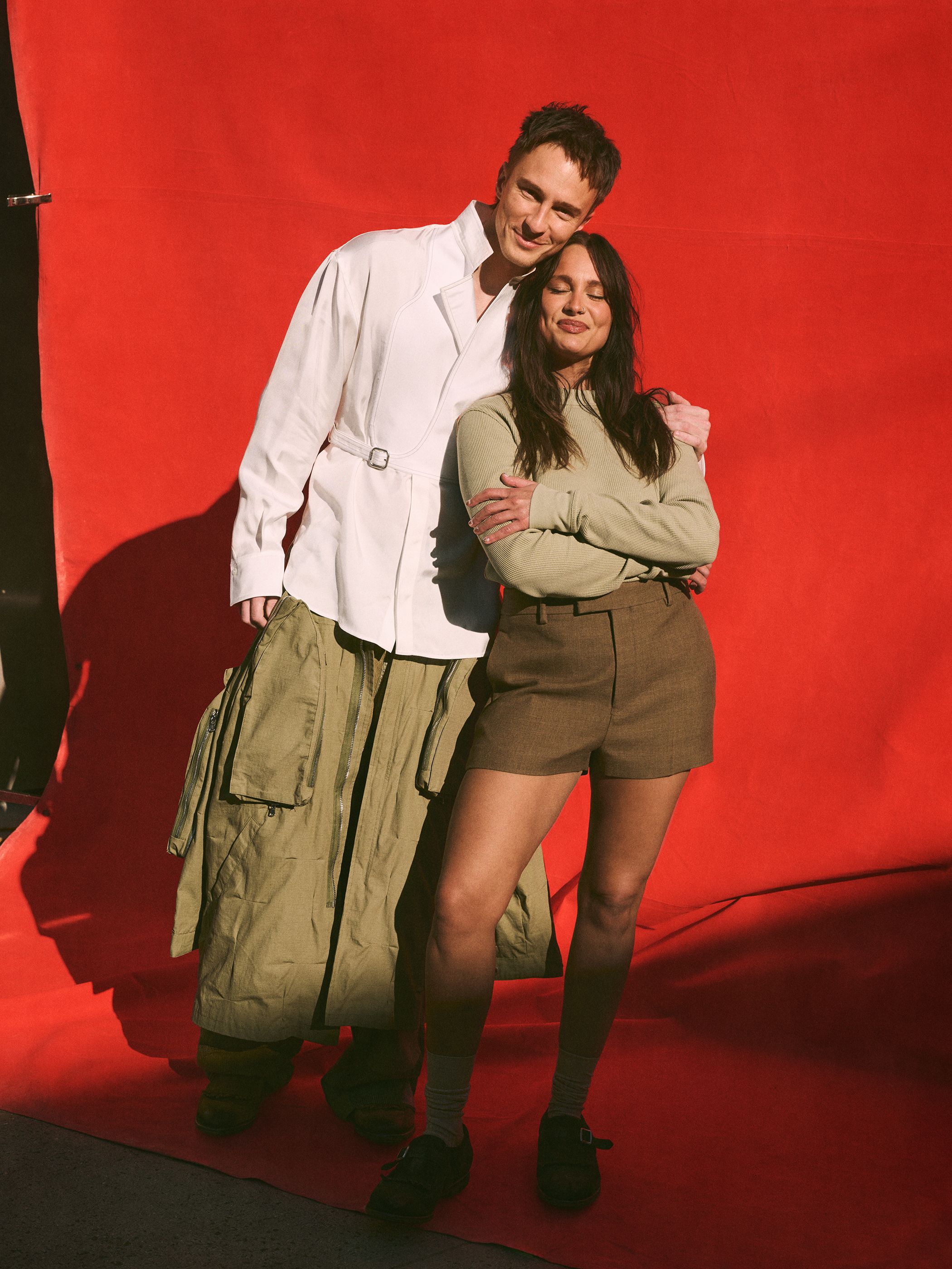 Studio shot of a stylish, smiling couple posing against a red background. The man in white, the woman in green sweater and brown shorts.