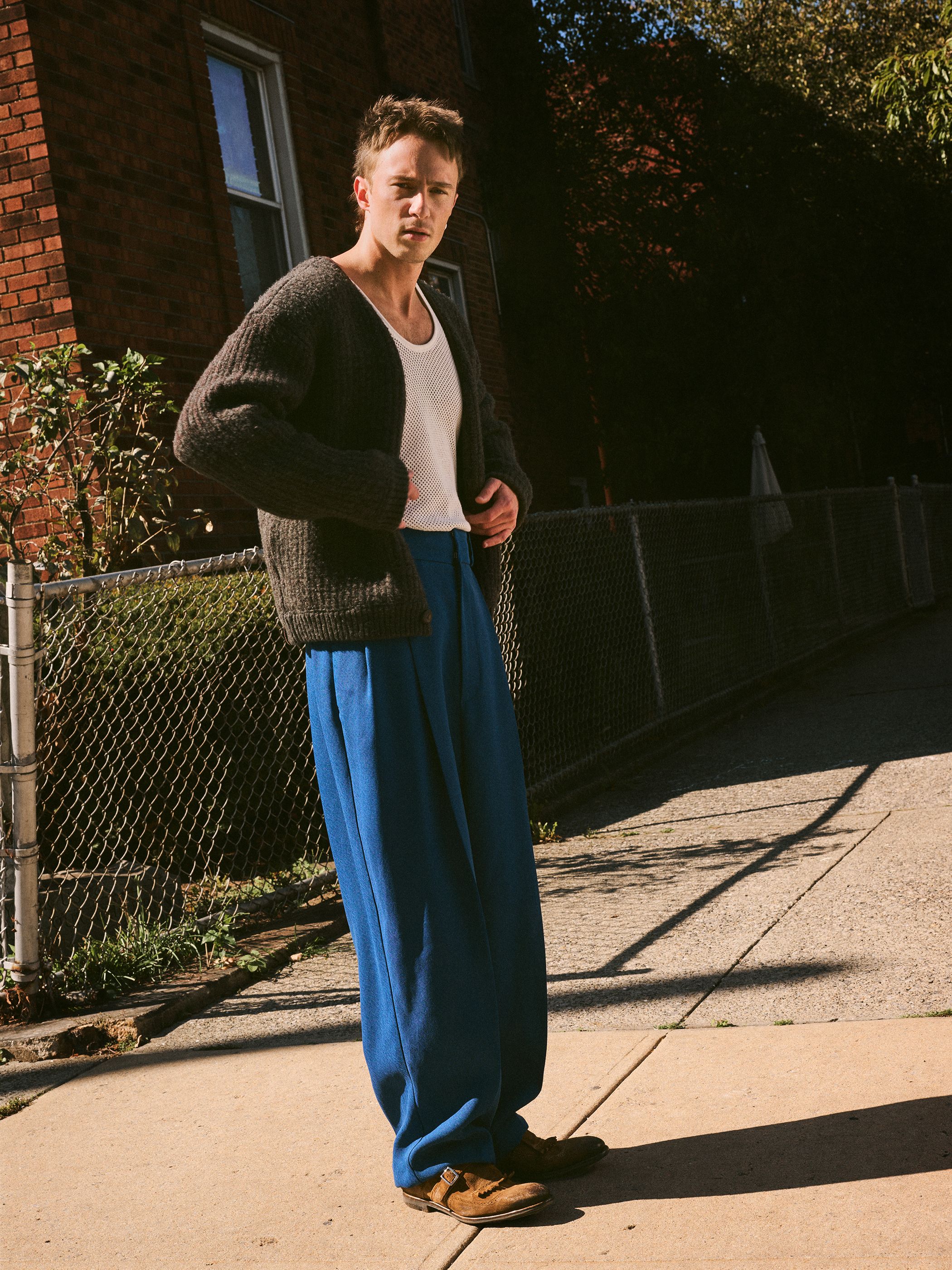 Adult male in a cardigan and blue pants stands on a sidewalk next to a brick building in an outdoor, eye-level medium shot.