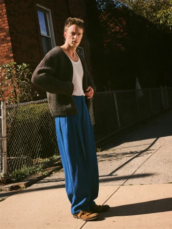 Adult male in a cardigan and blue pants stands on a sidewalk next to a brick building in an outdoor, eye-level medium shot.