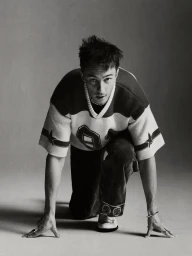 Drew Starkey crouching on a studio floor wearing a patterned athletic jersey and dark trousers against a neutral background.