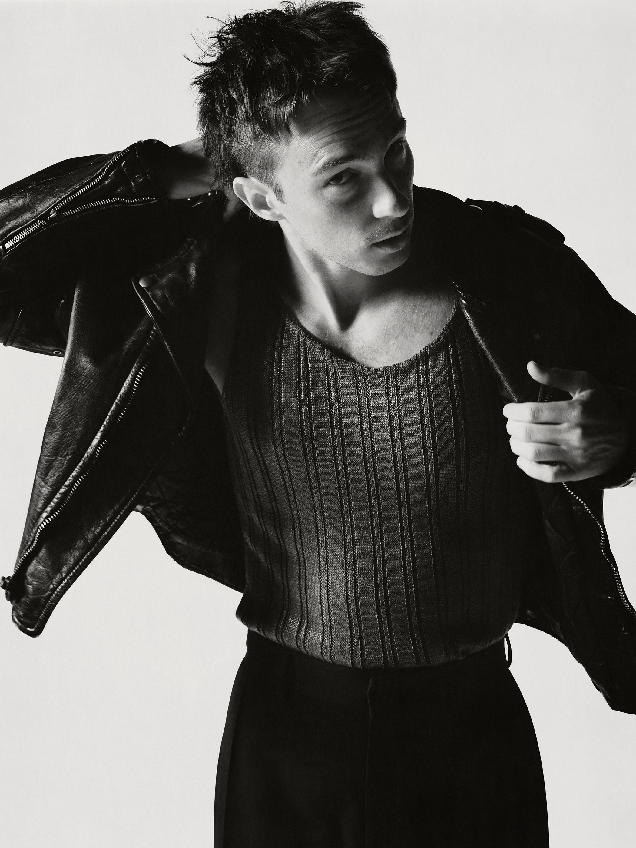 Monochrome portrait of a young adult man in a ribbed tank top and leather jacket against a white background in a studio setting.