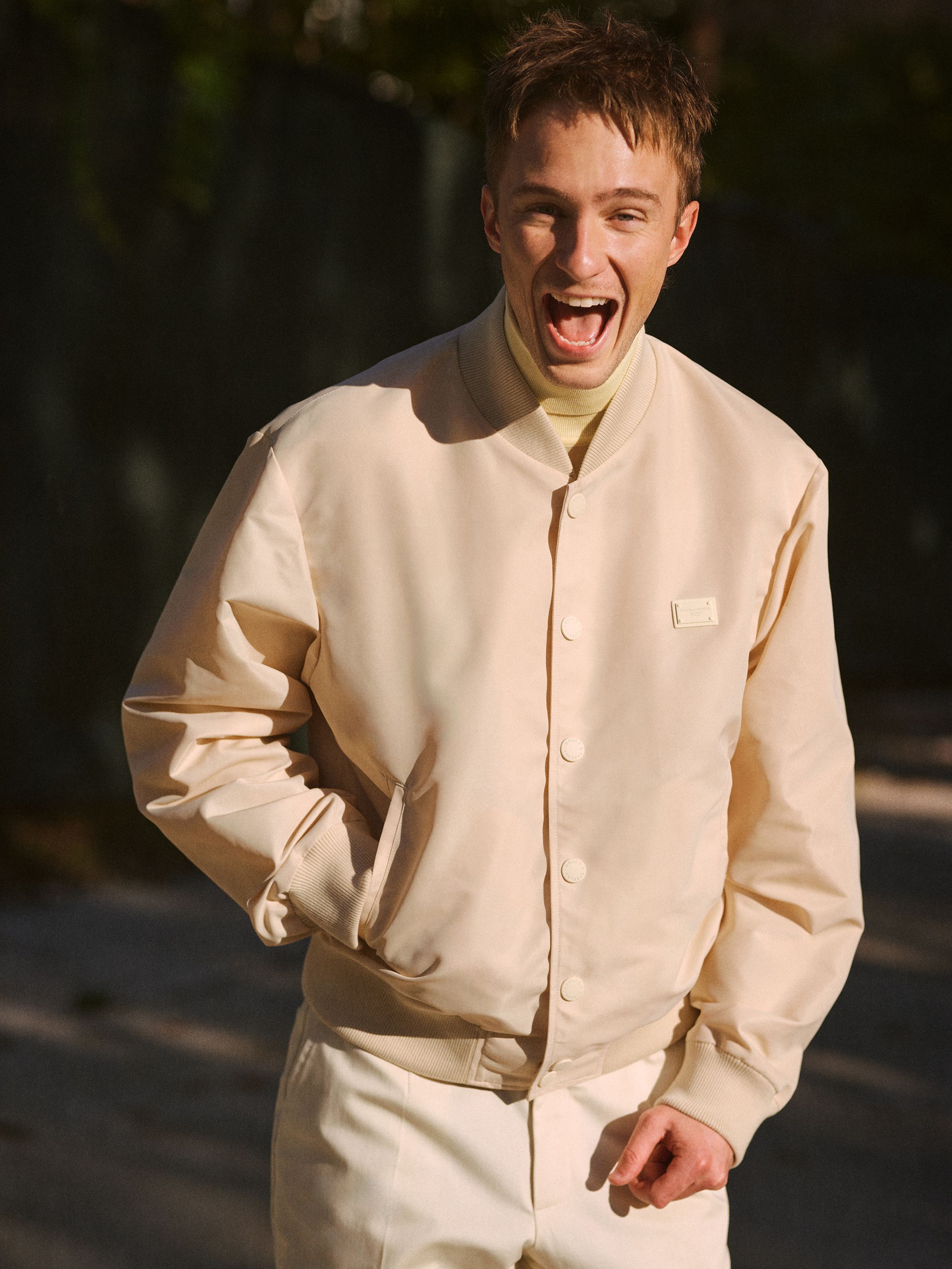 Young man smiles outdoors in an ivory bomber jacket, ecru pants, and yellow turtleneck, looking at the viewer with trees blurred behind him.