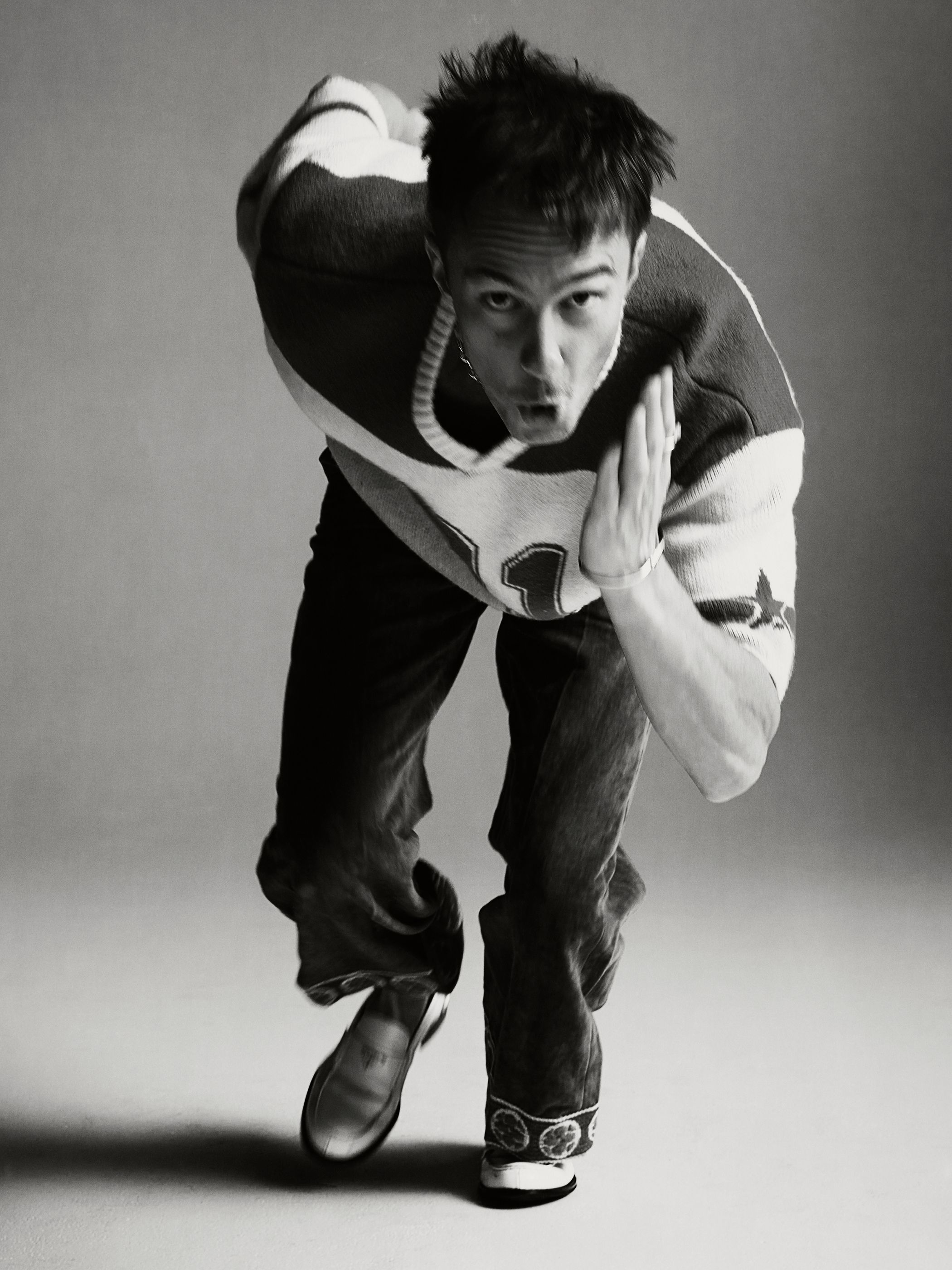 Black and white portrait of a man in motion, wearing a patterned shirt, jeans, and loafers, shot at eye-level in a photo studio.