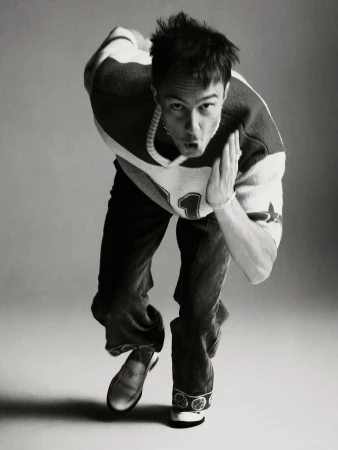 Black and white portrait of a man in motion, wearing a patterned shirt, jeans, and loafers, shot at eye-level in a photo studio.