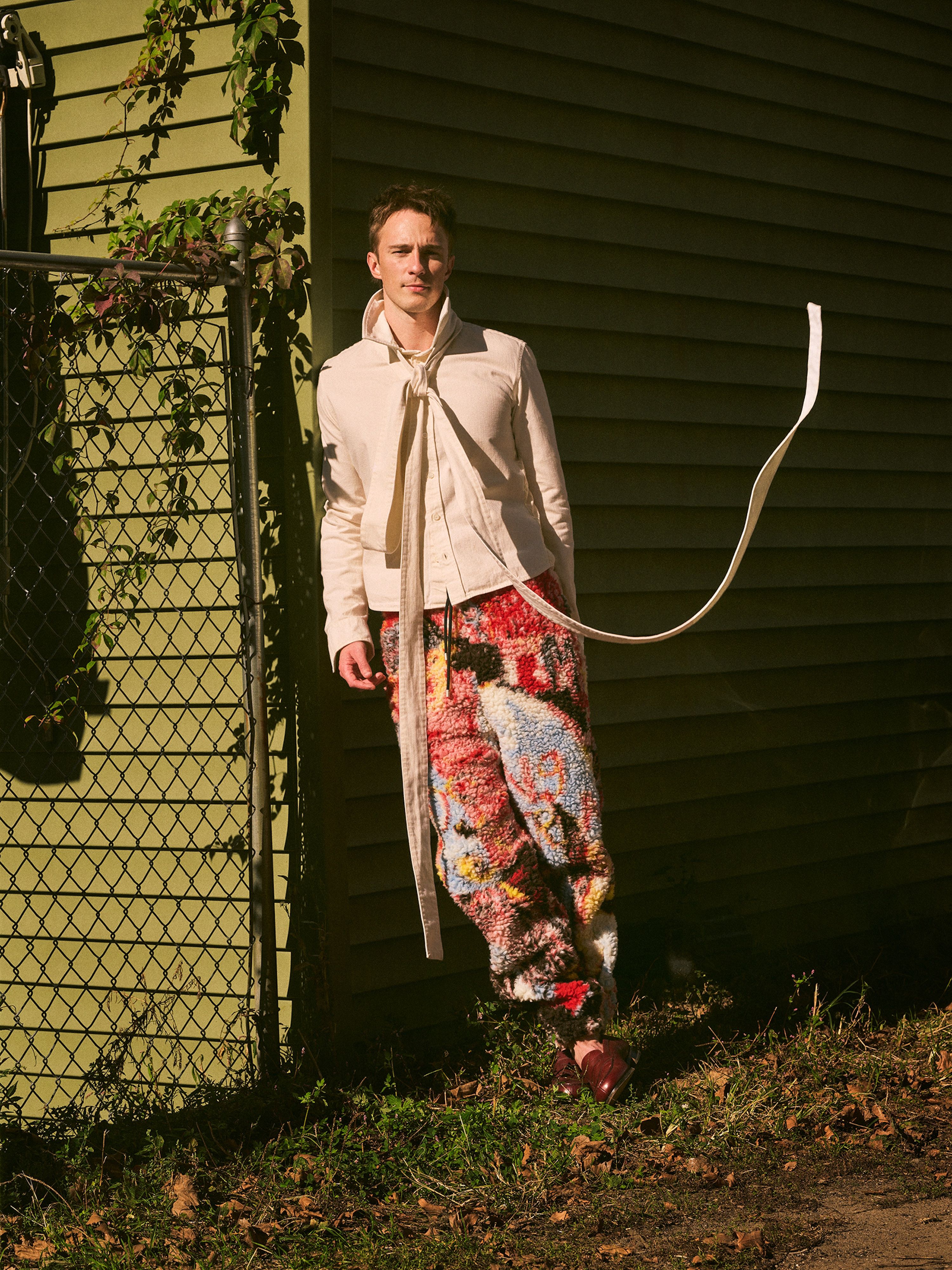 A man poses in front of a green building wearing a light jacket and colorful patterned pants, standing on grass in an outdoor environment.