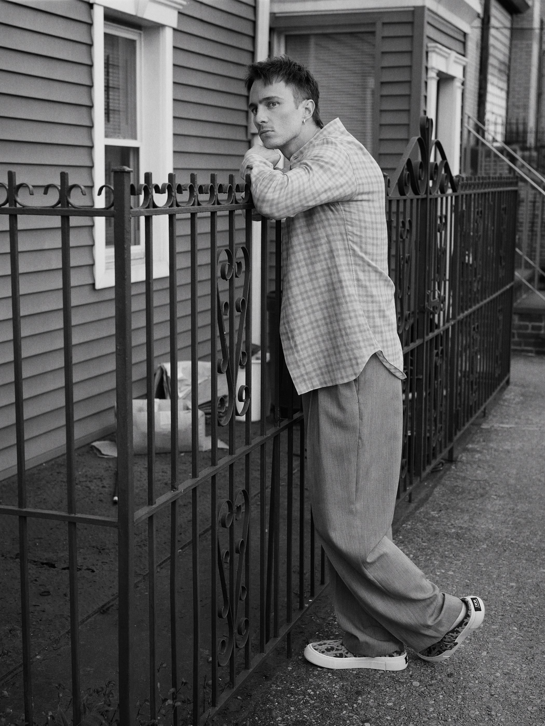 Black and white portrait of a young adult male leaning against a metal fence outside a house, wearing a button-down shirt and pants.