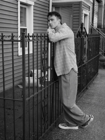 Black and white portrait of a young adult male leaning against a metal fence outside a house, wearing a button-down shirt and pants.