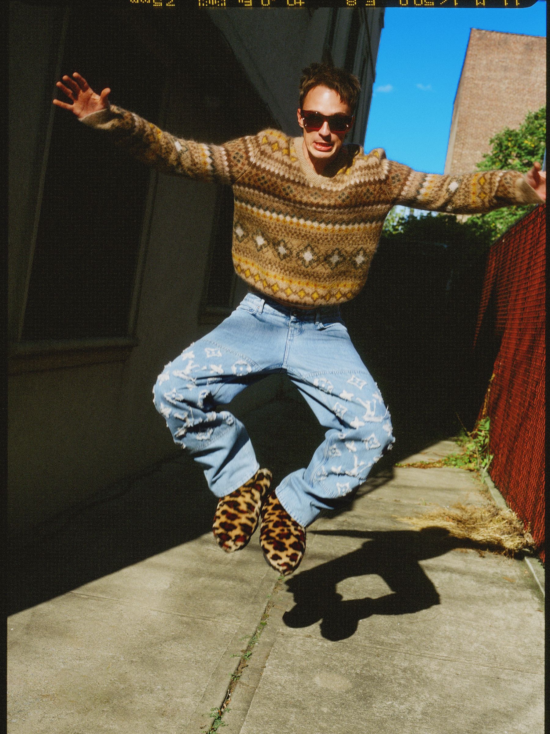 Man jumping in the air on a city street. He wears a patterned sweater, jeans, and leopard-print shoes against a blue sky backdrop.