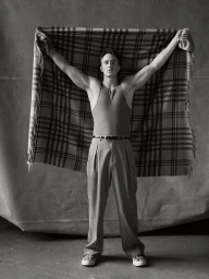 Drew Starkey holding a large plaid blanket behind him against a textured backdrop