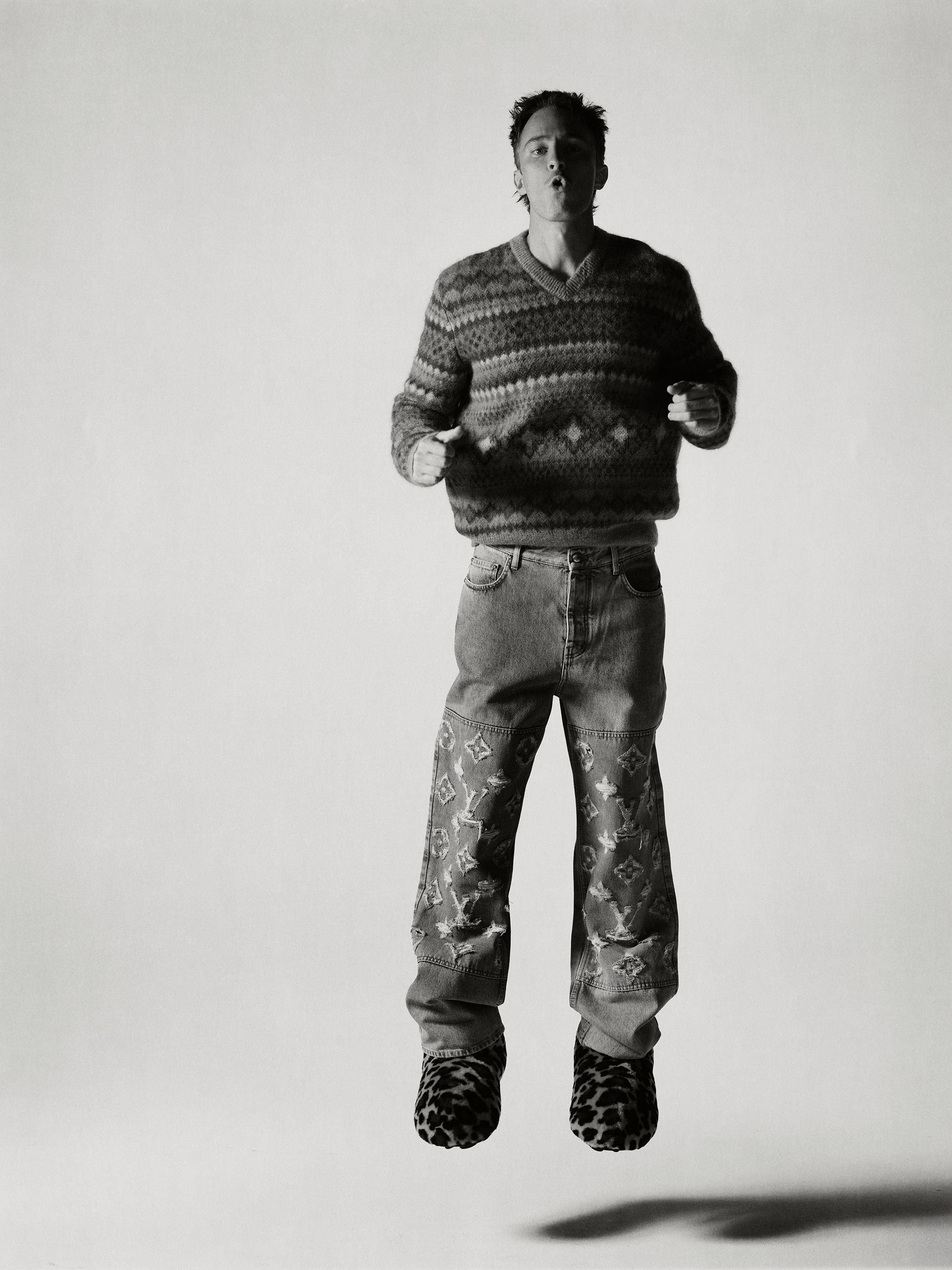 Drew Starkey jumping mid-air in a patterned sweater and patterned denim jeans against a light background
