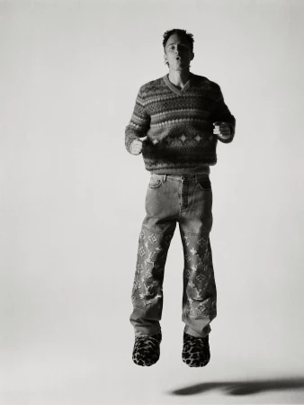 Drew Starkey jumping mid-air in a patterned sweater and patterned denim jeans against a light background