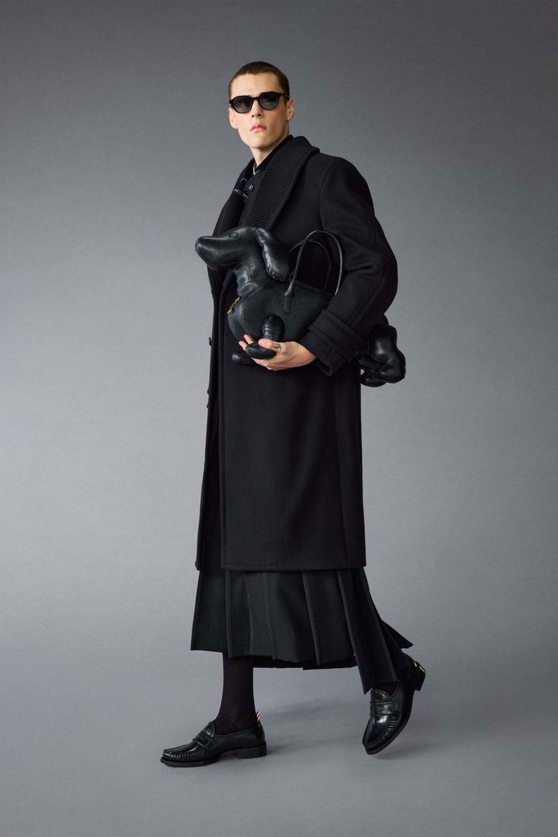 A stylish male fashion model in a long black coat holds a unique dachshund dog-shaped bag, complemented by sunglasses and shiny dress shoes.