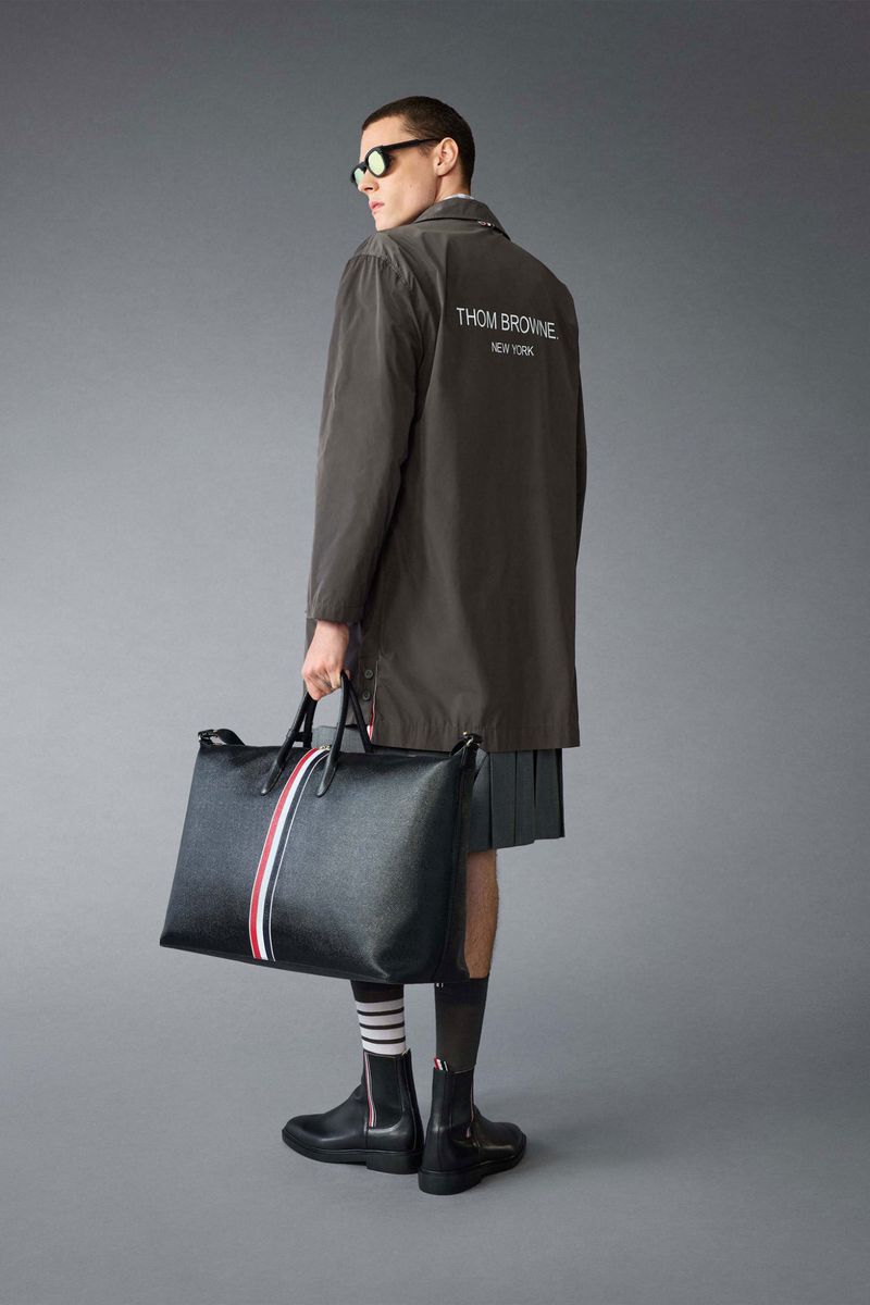 Man in olive coat and grey skirt carries a black leather bag with red, white, and blue stripe detail in front of a grey background.