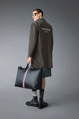 Man in olive coat and grey skirt carries a black leather bag with red, white, and blue stripe detail in front of a grey background.