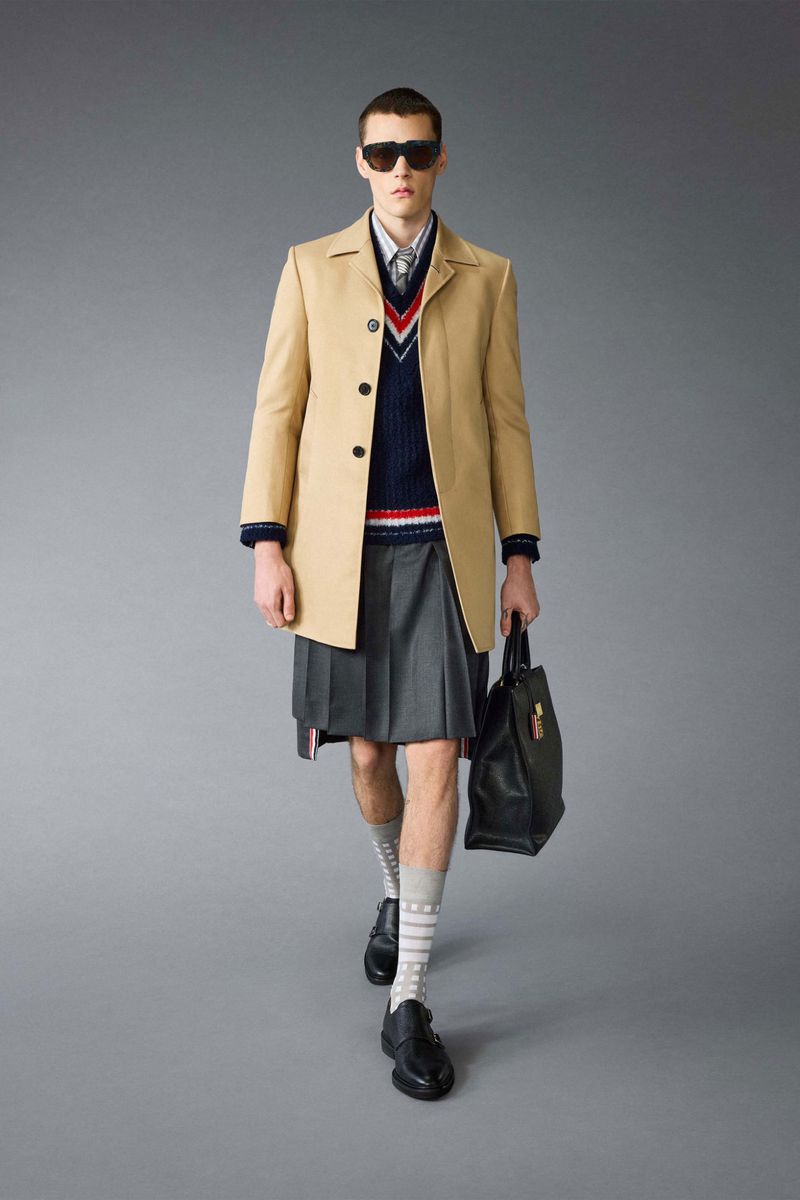 Male fashion model in coat, kilt, and sunglasses walking in studio with leather bag and patterned socks. Fashion editorial style.