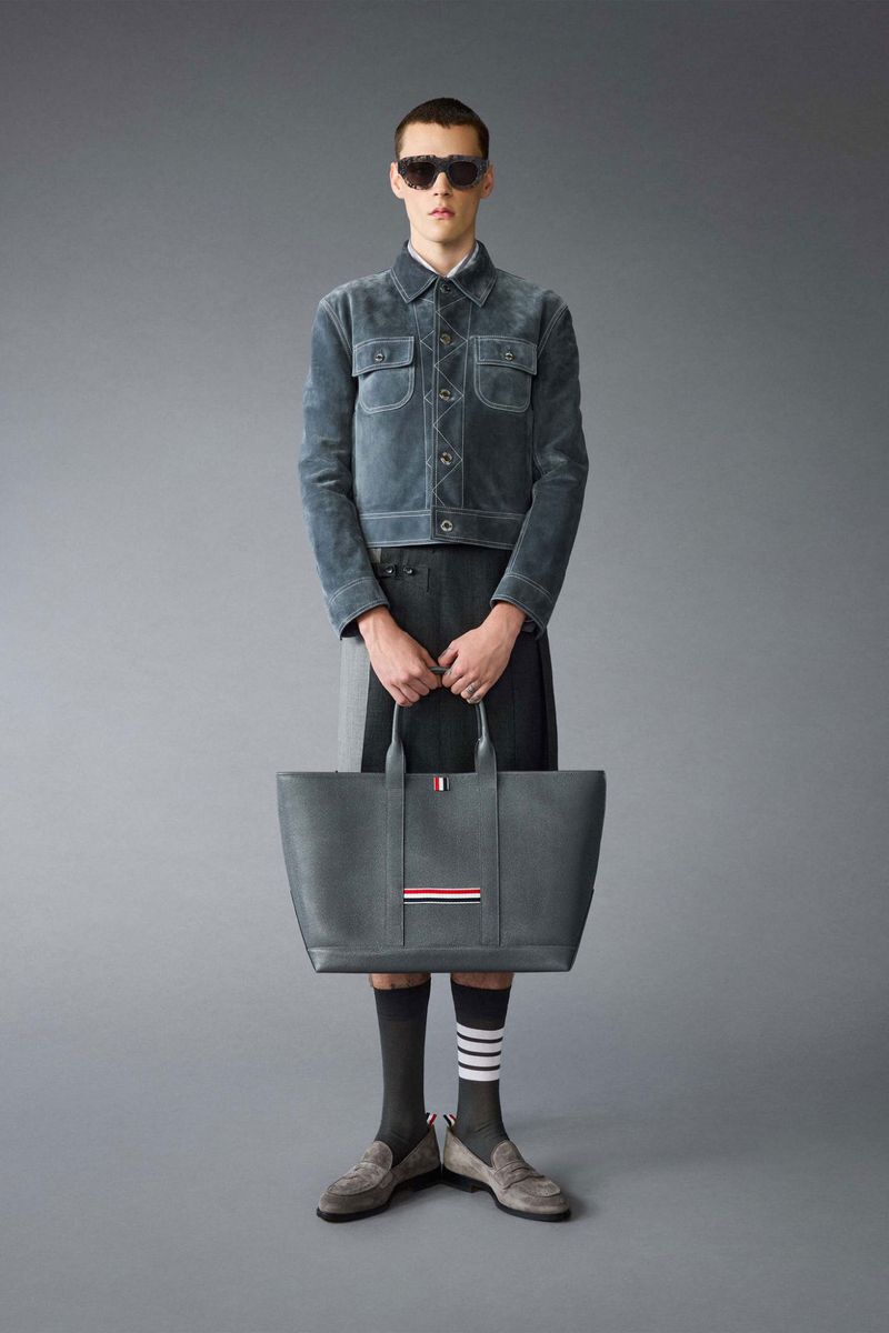 A slender male model poses in a studio wearing a blue jacket, grey skirt, and suede loafers, holding a grey tote bag with red stripes.