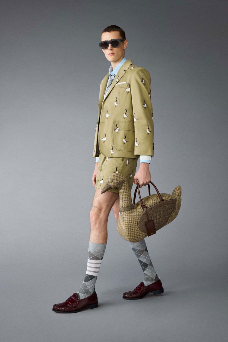 Fashion photo of a man in a tan suit with a goose pattern and sunglasses, holding a goose-shaped bag on a grey background.