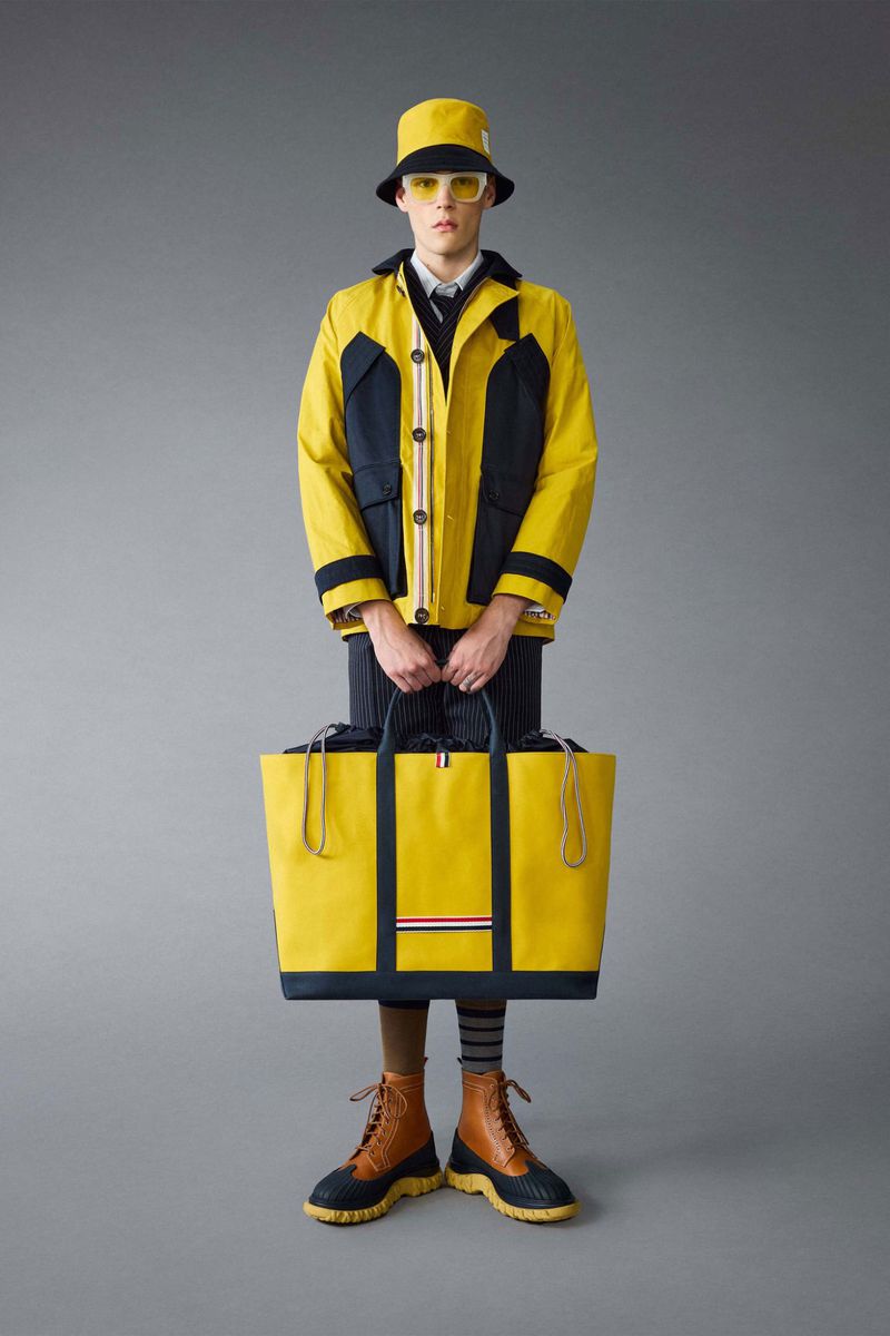 Fashion model in bucket hat and color-blocked jacket with large yellow tote bag against a grey background in a full-length shot.