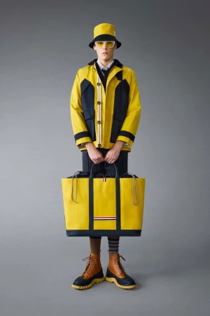 Fashion model in bucket hat and color-blocked jacket with large yellow tote bag against a grey background in a full-length shot.