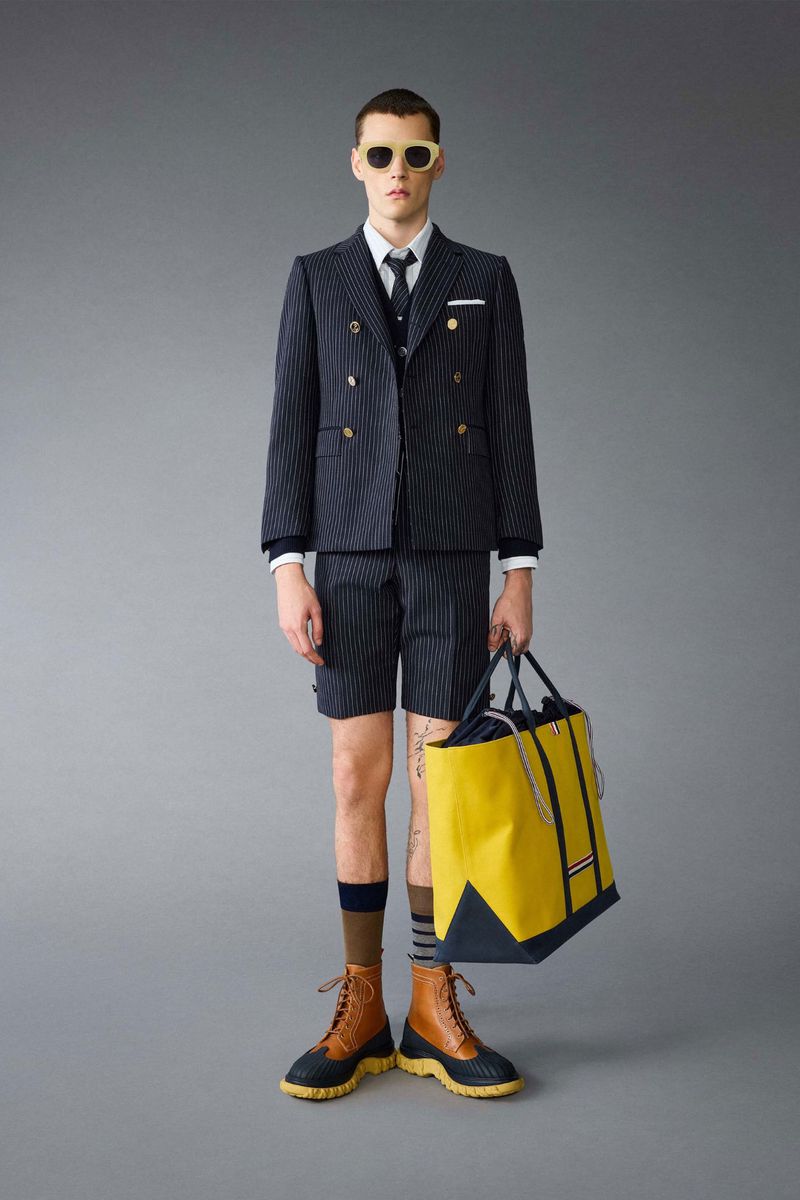 Man in pinstripe suit and sunglasses carrying a yellow tote bag, posing in a studio against a solid grey backdrop for fashion editorial.
